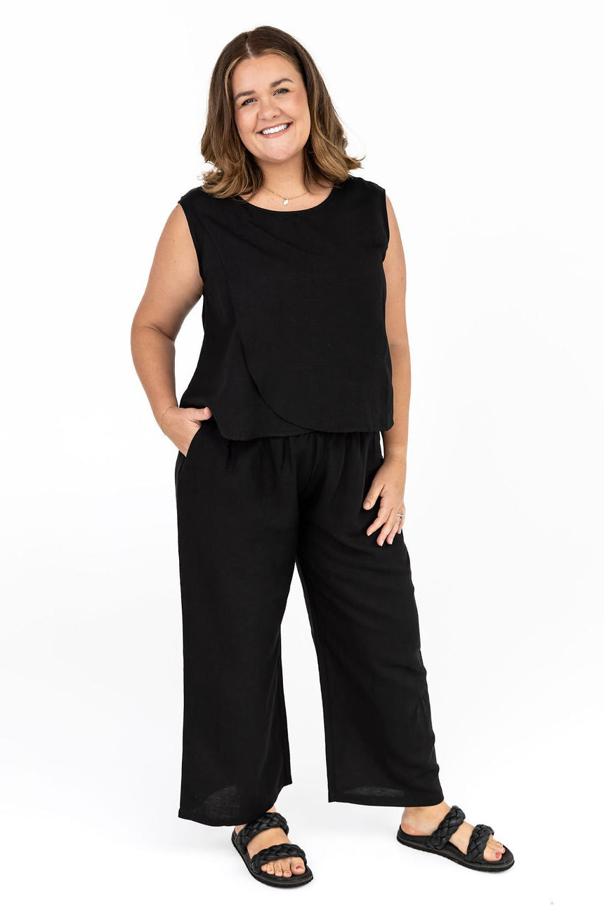 A woman standing, wearing a black linen-blend sleeveless top and wide-leg pants with a shirred waistband. She has dark hair and is smiling at the camera.