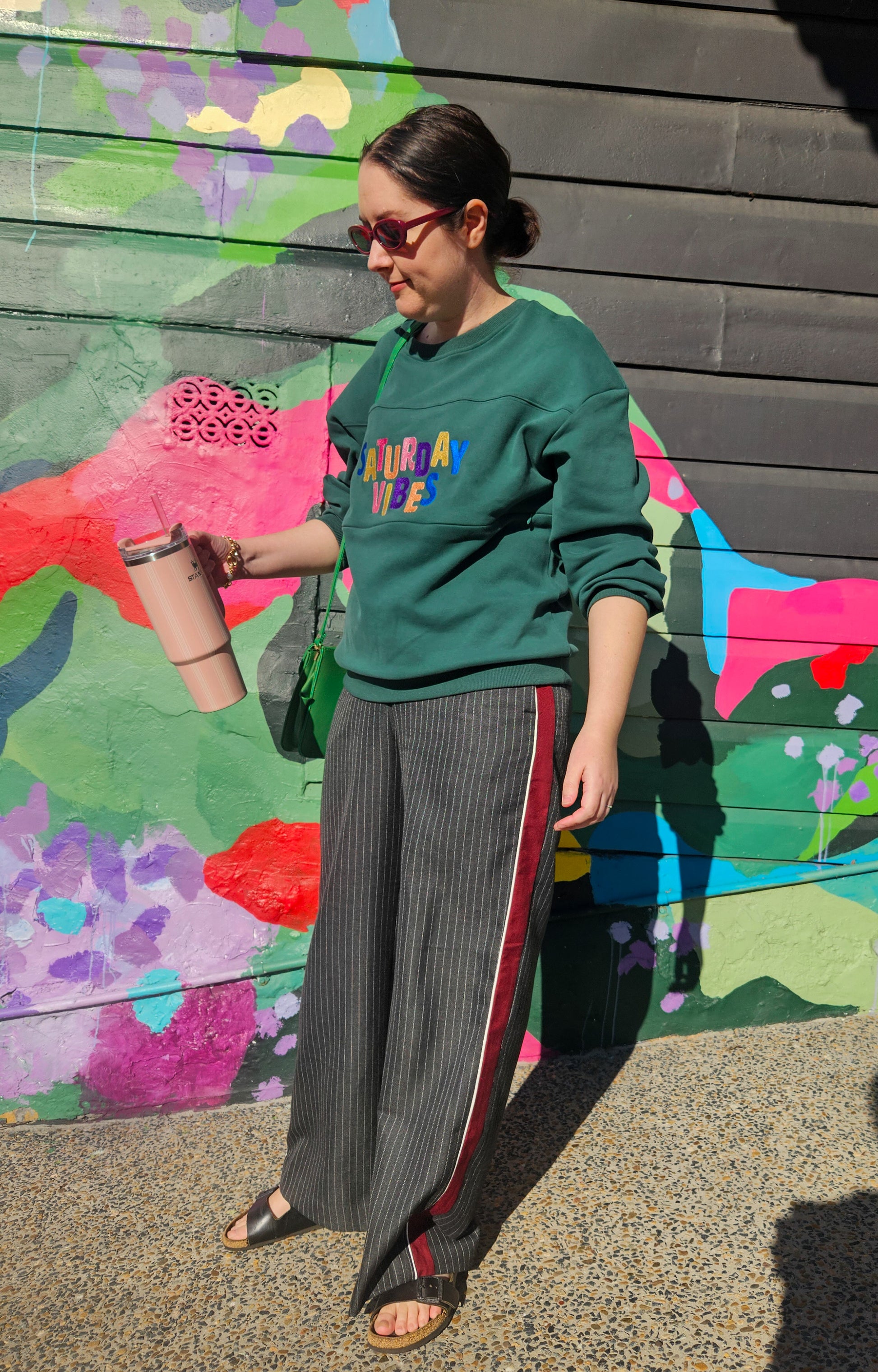 Person wearing a green sweatshirt with colorful text and gray pants with red stripes, standing in front of a colorful mural.
