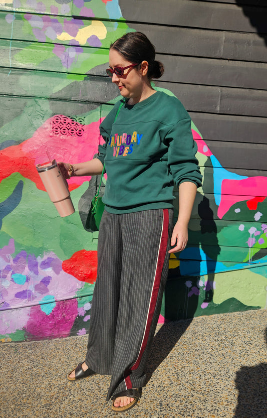 Person wearing a green sweatshirt with colorful text and gray pants with red stripes, standing in front of a colorful mural.